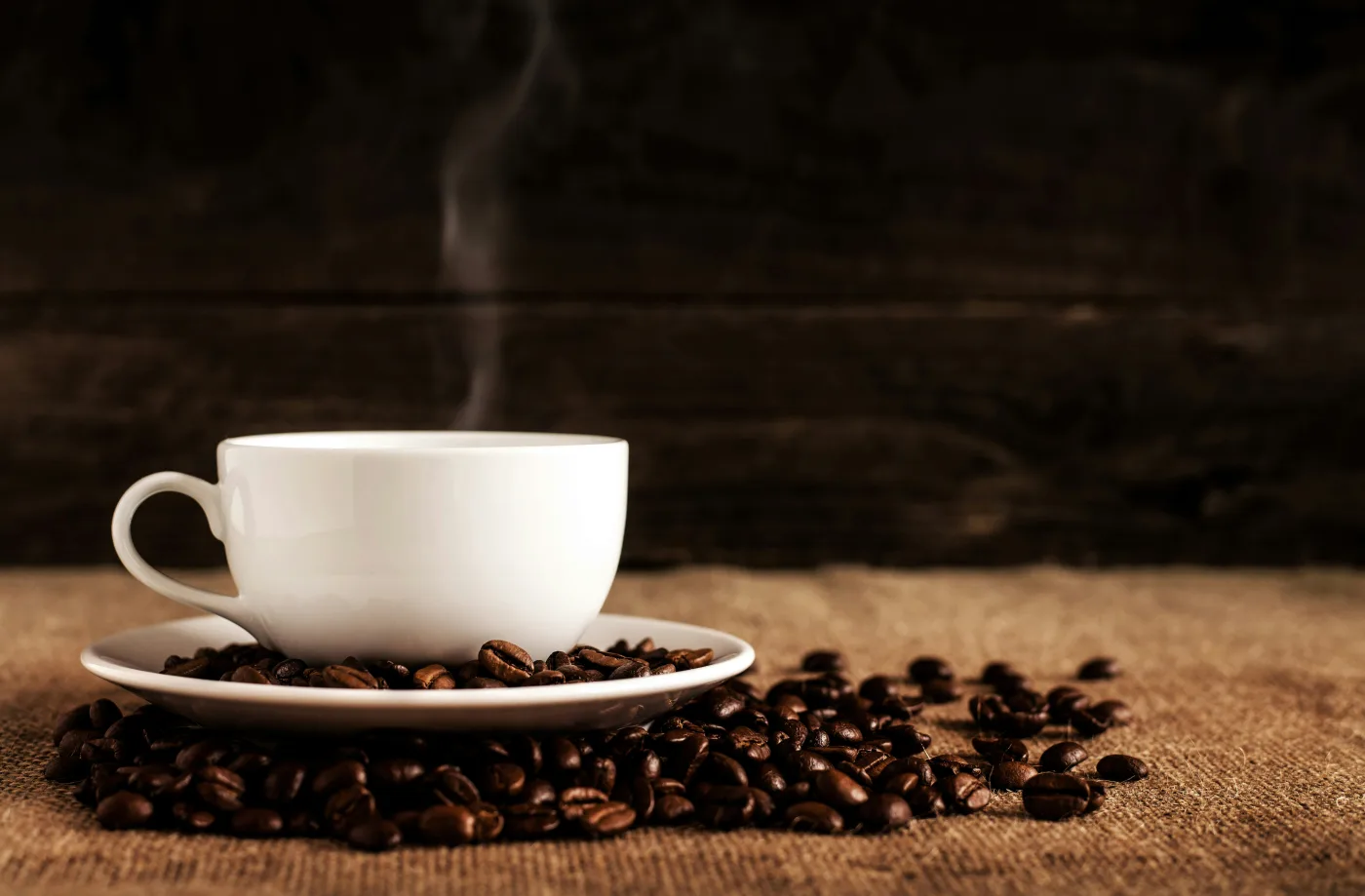 A freshly brewed cup of coffee, steam still rising, rests on a saucer surrounded by dark-roasted arabica beans on raw burlap — a scene repeated 1.5 billion times daily across the globe. New research from Queen Mary University of London suggests that every sip may be activating AMPK, an ancient cellular energy sensor tied to stress resilience and longer lifespan. Image credit: NavsoraTimes / Licens