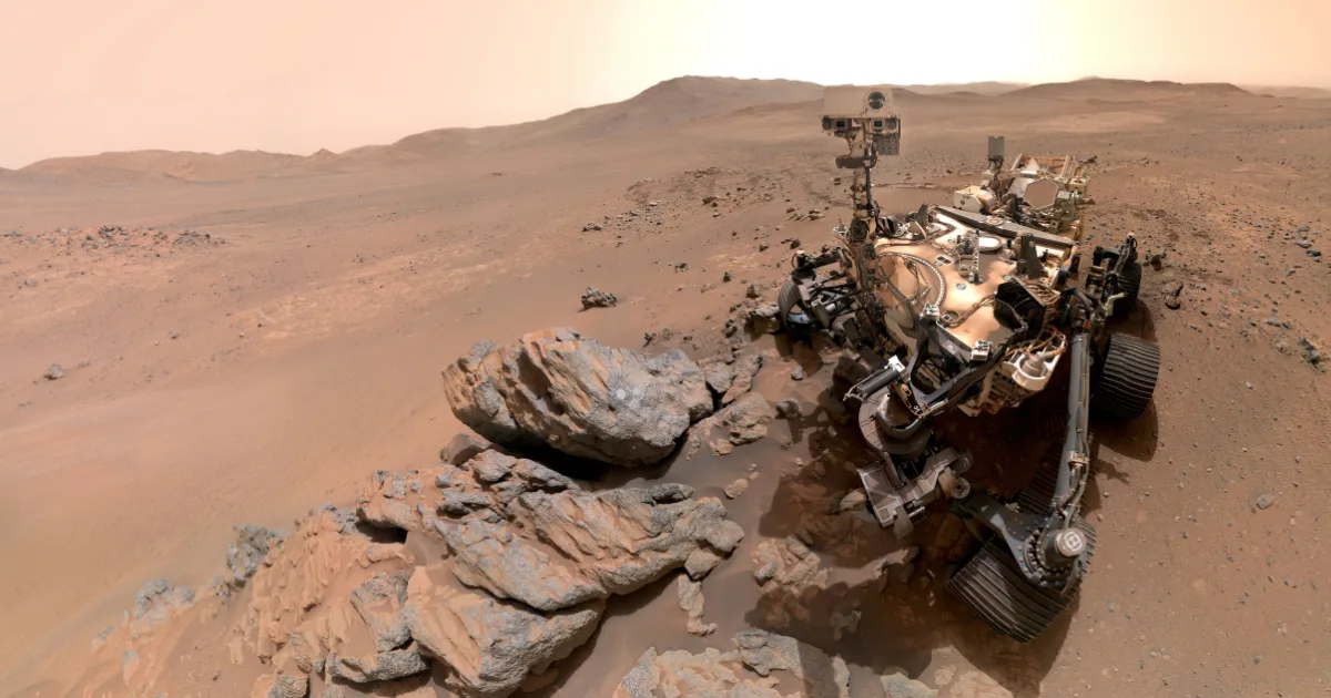 NASA's Perseverance rover peers down at the rocky "Arethusa" outcrop before turning its camera toward the viewer, in a 61-image composite captured at the farthest west point of the mission. The orange-red terrain behind it belongs to territory never before visited by any rover. Image: NASA/JPL-Caltech/MSSS.