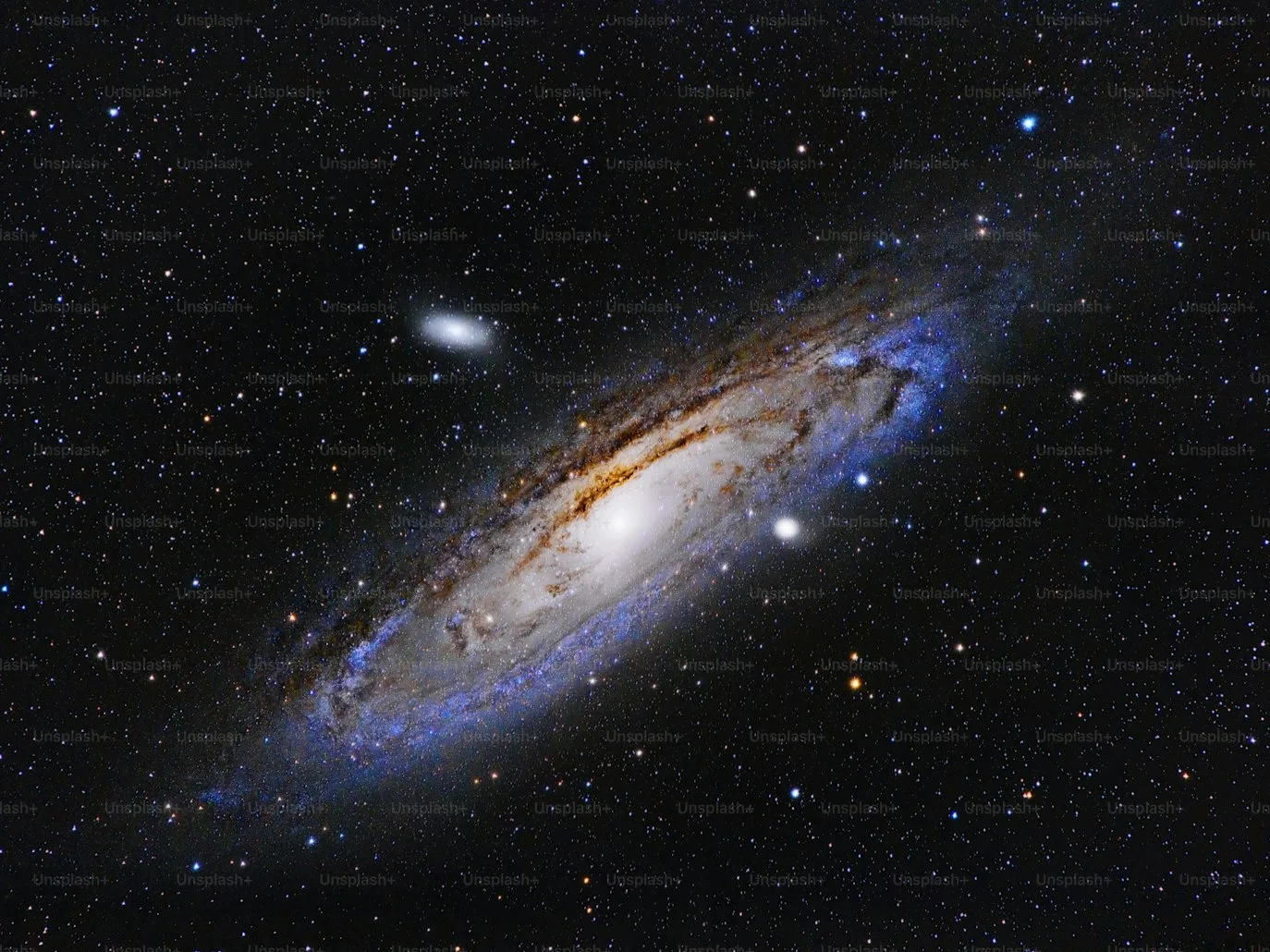 The Andromeda Galaxy stretches across this deep-sky photograph, its blazing white core fading into golden dust lanes and electric-blue spiral arms packed with young stars. The small elliptical object in the upper left is M32, one of Andromeda's satellite galaxies. Along with the Milky Way and about 80 other dwarf galaxies, both of these objects are members of the Local Group — the galactic family